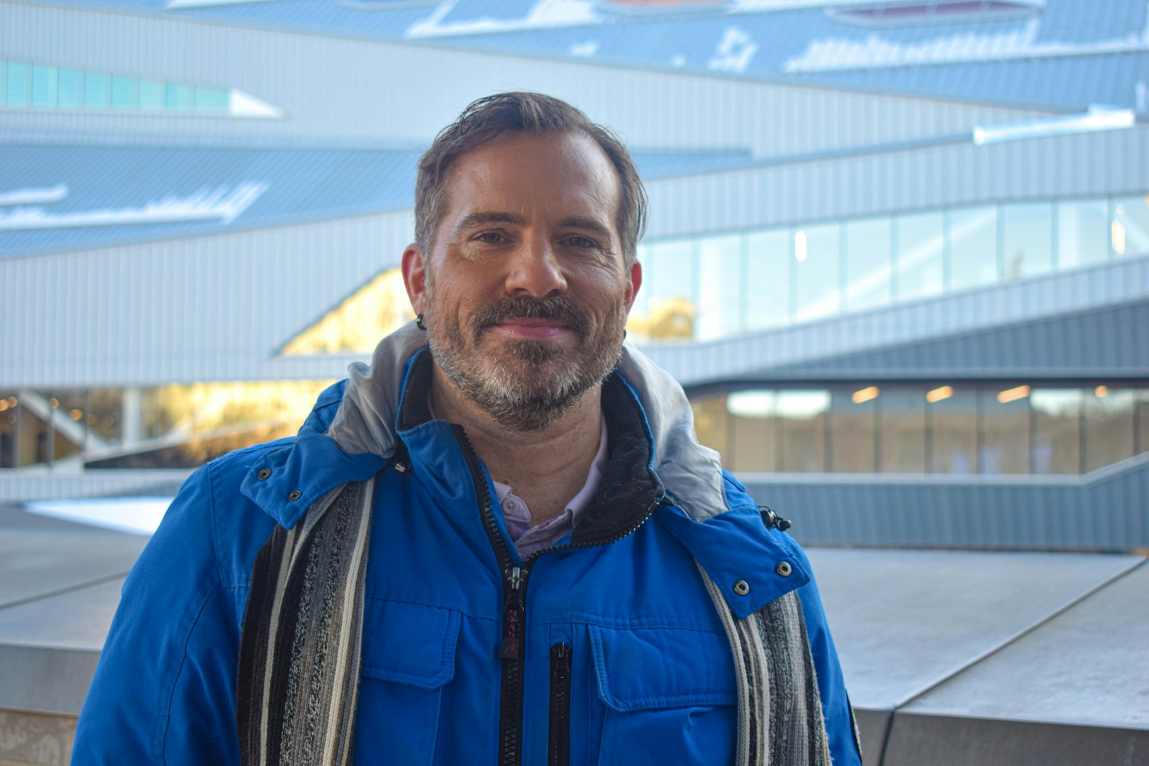 A man with medium-length wavy hair, wearing a teal work jacket, stands looking directly at the camera with a serious expression. The background features blurred white containers, suggesting a workplace environment.