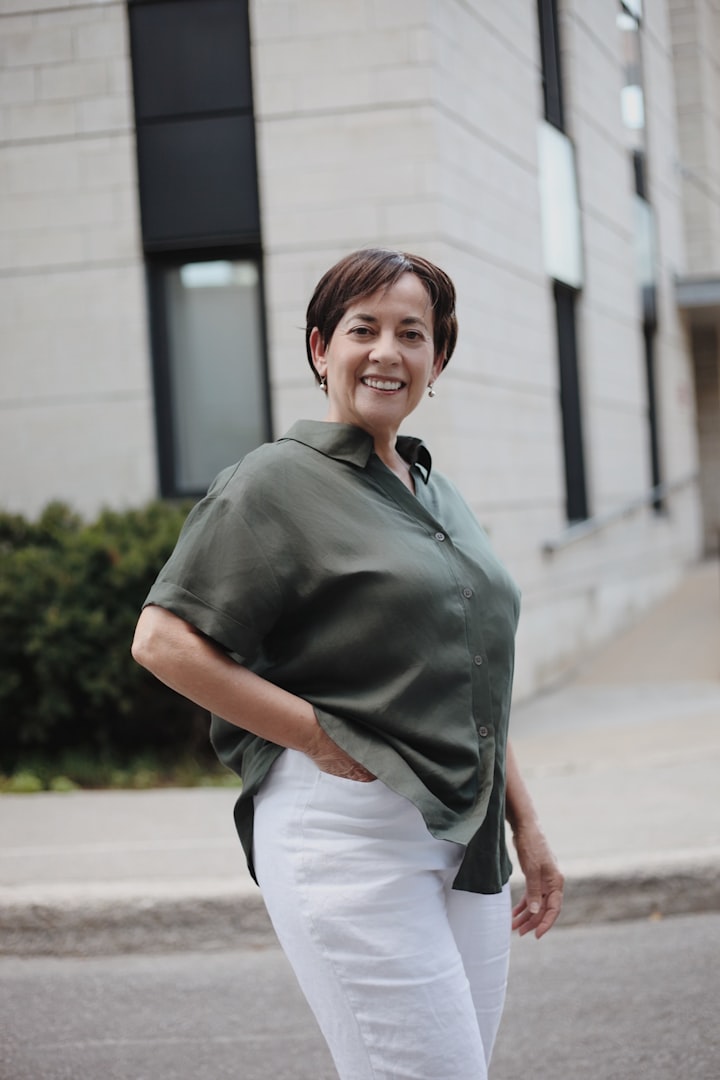 A woman with wavy, shoulder-length brown hair is smiling gently. She is wearing a white blouse and is standing in a bright, minimalist setting with turquoise decorative items in the background.
